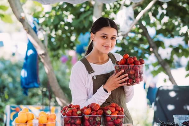Young positive salesgirl at job, selling sells home-grown vegetables and fruits in vegetable shop - Fotografie, Obrázek