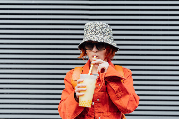 Hipster fashion young woman in bright clothes and bucket hat drinking fruity sugar flavored tapioca pearl bubble tea with straw on the gray striped wall background. Selective focus. Copy space - Photo, image
