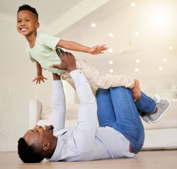 Adorable little african american boy playing in the living room pretending to fly at home with his father. Cute black little male child smiling and bonding with his dad inside. Man having so much fun. - 写真・画像
