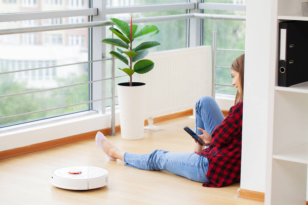 White robotic vacuum cleaner cleaning the floor while woman sitting near wall and rest - Φωτογραφία, εικόνα