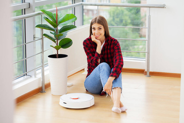 White robotic vacuum cleaner cleaning the floor while woman sitting near wall and rest - Photo, Image
