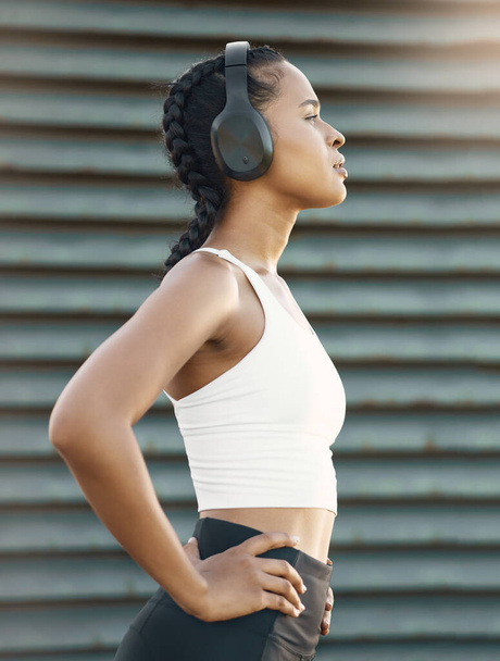 One fit young hispanic woman wearing headphones and taking a rest break to catch her breath after a run or jog in an urban setting outdoors. Female athlete looking tired after intense cardio exercise. - Foto, Bild