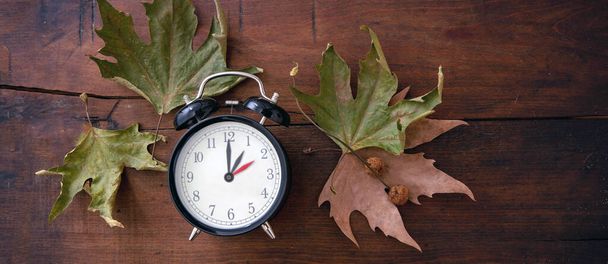 Fall Back, Daylight Saving Time. One hour back on yellow clock and autumn leaves on wooden background, top vie - Photo, Image