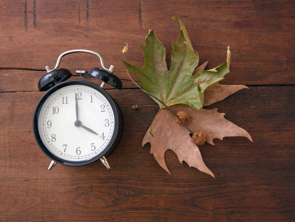 Υποχώρηση, θερινή ώρα. Μαύρο ξυπνητήρι και φύλλα του φθινοπώρου σε ξύλινο τραπέζι, κορυφή vie - Φωτογραφία, εικόνα