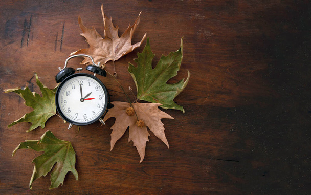 Fall Back, Daylight Saving Time. One hour back on yellow clock and autumn leaves on wooden background, top vie - Photo, Image