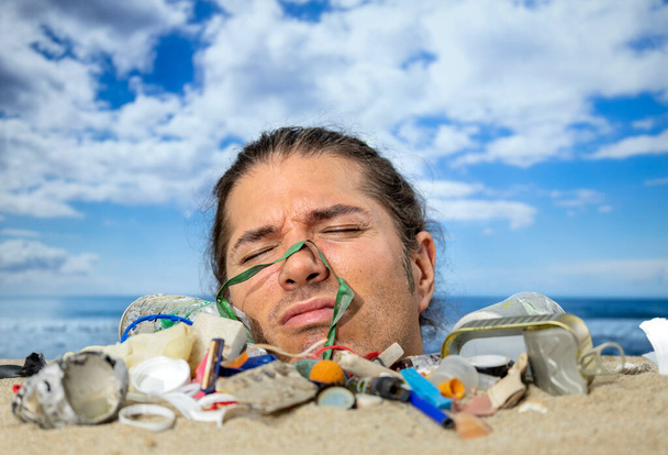 Un uomo sepolto fino alla testa su una spiaggia coperta di spazzatura e rifiuti di plastica in movimento  - Foto, immagini