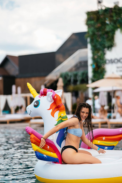 Woman in bikini in inflatable unicorn toy mattress float in pool. Girl relaxing sunbathing enjoying travel holidays at resort pool - Fotó, kép