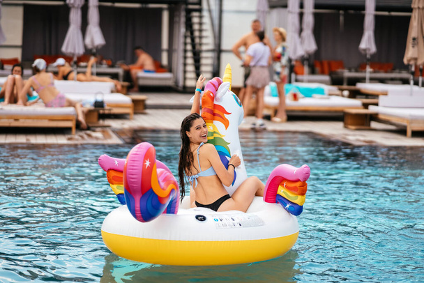 Γυναίκα με μπικίνι σε φουσκωτό στρώμα παιχνιδιού με μονόκερο επιπλέει στην πισίνα. Κορίτσι χαλαρωτική ηλιοθεραπεία απολαμβάνοντας ταξιδιωτικές διακοπές στην πισίνα θέρετρο - Φωτογραφία, εικόνα