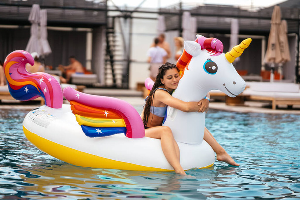 Женщина в бикини в надувном однорогом игрушечном матраце плавает в бассейне. Девушка расслабляется загорать наслаждаясь отпуском в курортном бассейне - Фото, изображение