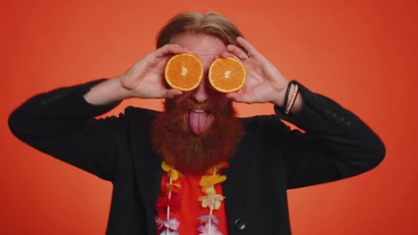 Όμορφος νεαρός τουρίστας βάζοντας τα μισά πορτοκάλια στα μάτια, χορτοφαγικός τρόπος ζωής, βιταμίνες για την υγεία, καλές διακοπές, βιολογικά οικολογικά τρόφιμα. Νεαρέ αγόρι απομονωμένο στο φόντο του πορτοκαλί στούντιο - Πλάνα, βίντεο