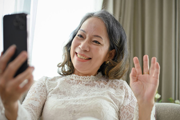 Attraente e felice asiatica donna di mezza età utilizza il suo smartphone per videochiamare, agitando la mano per salutare e salutare la sua amica nel suo soggiorno. - Foto, immagini