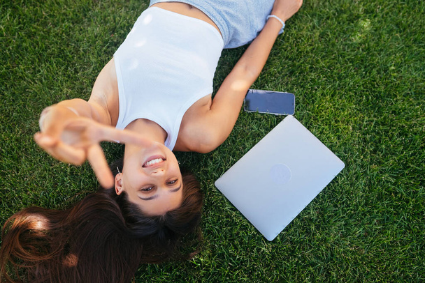 Young fit attractive woman lying on green lawn park. Perks of freelance concept. Background, copy space - 写真・画像