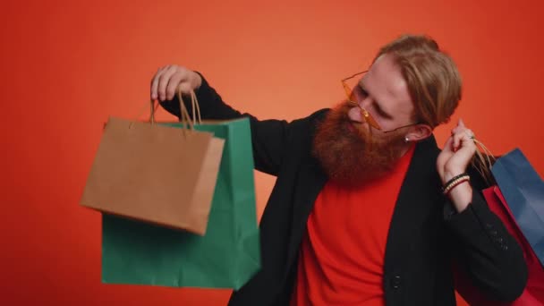Счастливый турист в черной куртке, показывающий сумки, рекламные скидки, улыбающийся взгляд поражен низкими ценами, шоппинг в Черную пятницу праздников. Молодой бородатый парень на оранжевом фоне - Кадры, видео