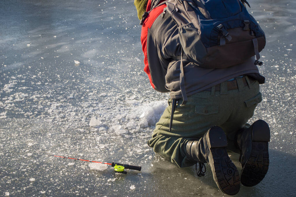 Un hombre en una chaqueta de invierno, pantalones y con una mochila y girando cerca perfora diligentemente el hielo en el río congelado con un hacha. Vista trasera - Foto, Imagen