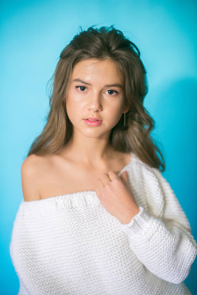 A cute teenage girl with long dark hair in trendy jeans and a white knitted sweater on a blue background in the studio. Fashion for teenagers - Fotografie, Obrázek