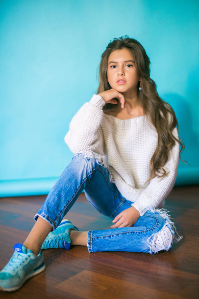 A cute teenage girl with long dark hair in trendy jeans and a white knitted sweater on a blue background in the studio. Fashion for teenagers - Foto, Imagen