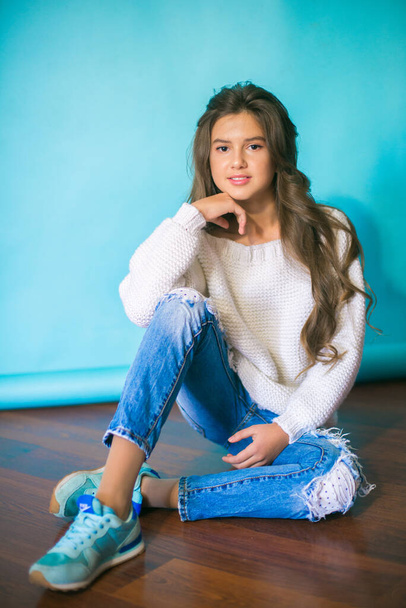 A cute teenage girl with long dark hair in trendy jeans and a white knitted sweater on a blue background in the studio. Fashion for teenagers - Fotó, kép