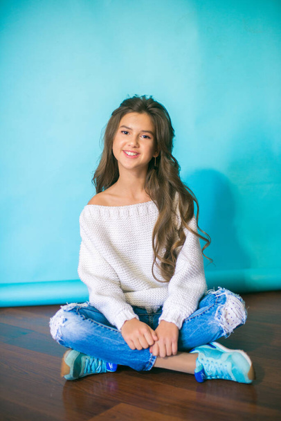 A cute teenage girl with long dark hair in trendy jeans and a white knitted sweater on a blue background in the studio. Fashion for teenagers - Foto, Imagem