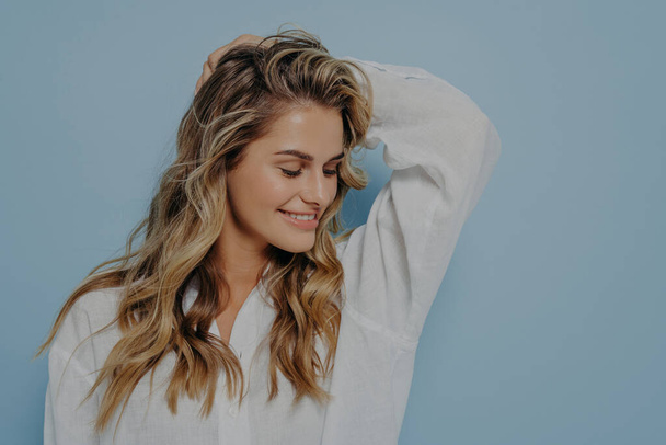 Relaxed romantic blonde woman with long wavy hair and hand behind her head, looking down with bright smile, dressed in white shirt with unbuttoned top while standing alone next to light blue wall - Photo, Image