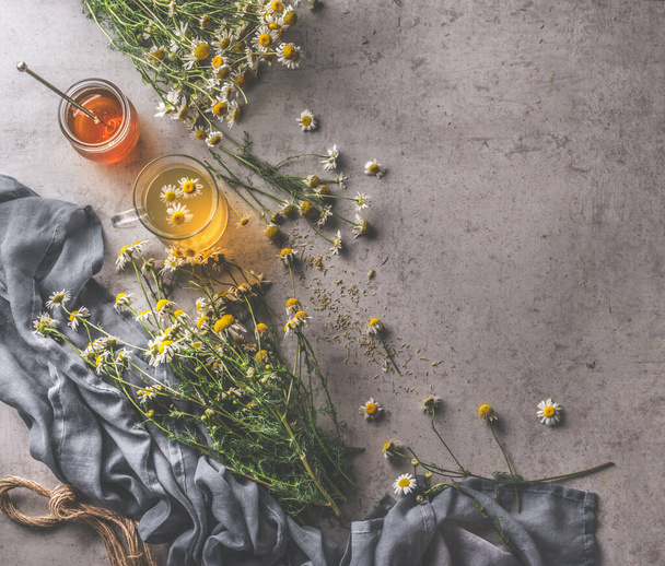 Fresh chamomile tea in glass cup, honey and honey spoon, medicinal plants. Natural treatment with herbs. Dark cloth and cord on dark concrete background. Top view. - Foto, imagen