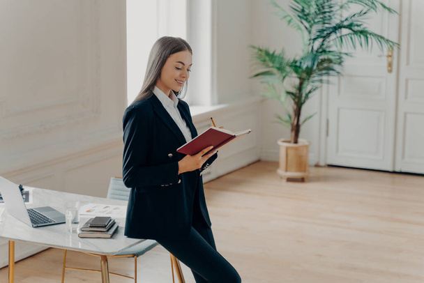 Side portrait of attractive business woman in elegant stylish outfit planning working day, making notes in notebook while leaning on white desk in big office with palm tree in pot. People at work - Photo, Image