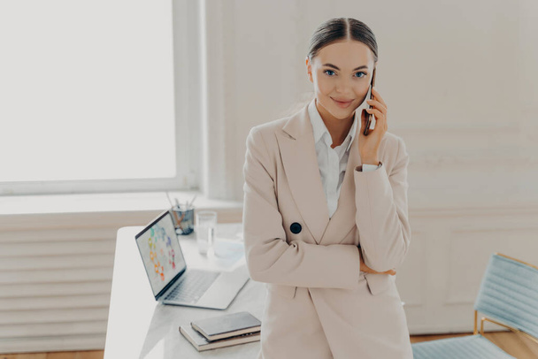 明るい色のオフィスで働く正式なスーツのビジネスマンの女性は、携帯電話で話をし、カメラを喜んで見て。女性監督でベージュ形式的なスーツの考え方の肯定的な結果の会話 - 写真・画像
