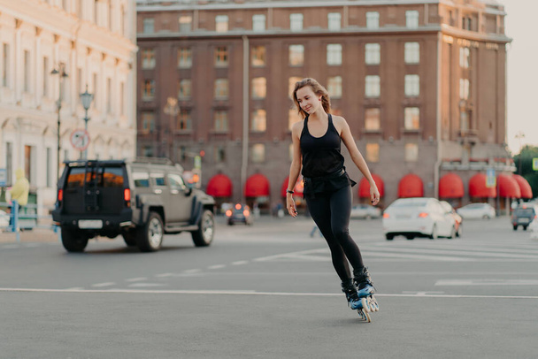 Active girl rollerblading on grey asphalt poses on rollers dressed in active wear drives fast leads healty lifestyle enjoys favorite hobby. Sport recreation roller skating summer time concept - Photo, Image