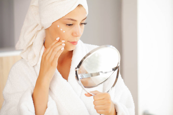 Γυναίκα με πετσέτα στο κεφάλι στο μπάνιο κοιτάζει και αγγίζει το πρόσωπό της στον καθρέφτη και απολαμβάνει τη νιότη και την ενυδάτωση - Φωτογραφία, εικόνα