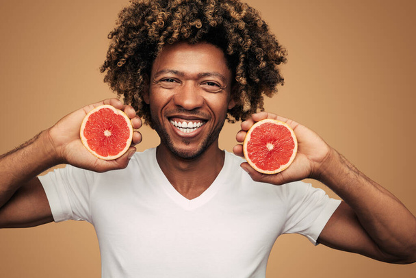 Optimistic young African American male model with curly dark hair in white t shirt, smiling brightly and looking at camera while demonstrating halved grapefruit against brown background - Photo, image