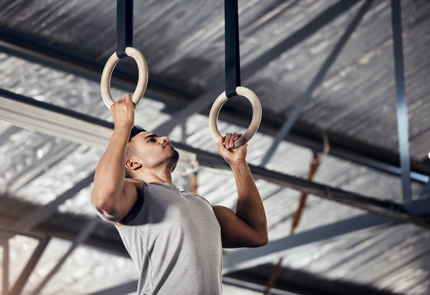 Fitness, exercise man in gymnastics gym during pull up, training or exercise on rings. Young sports athlete or gymnast, strong mindset and start workout to improve muscle or cardio at sport club. - 写真・画像
