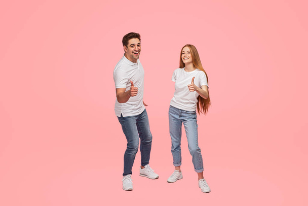 Full body of delighted young couple in similar jeans and white t shirts, smiling and looking at camera while showing thumbs up gesture against pink background - Foto, Imagen
