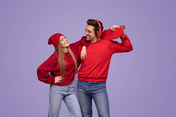 Positive young enamored boyfriend and girlfriend in in red sweatshirts hat and headphones, smiling and looking at each other while standing against purple background with skateboard - Photo, Image