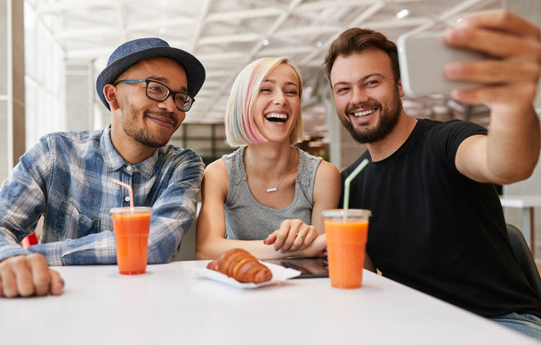 Cheerful young multiracial male and female best friends in casual clothes, smiling happily while taking selfie on smartphone sitting at table with juices and croissant in cafe - Photo, Image