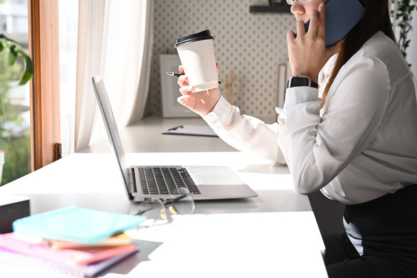 Sivunäkymä nuori nainen toimistotyöntekijä juo kahvia ja puhuu matkapuhelimella istuessaan työpaikallaan. - Valokuva, kuva