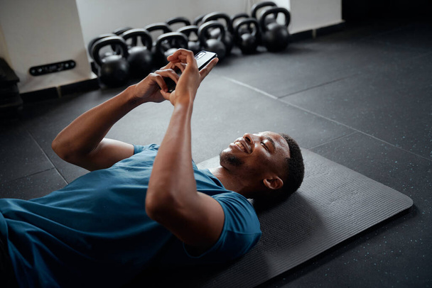 Young black man in sportswear typing on mobile phone while resting at the gym - 写真・画像