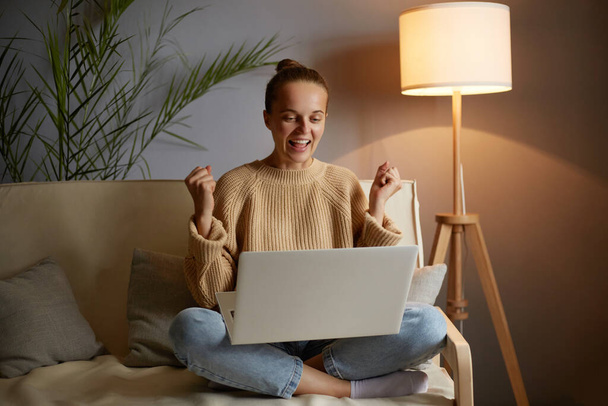 Indoor shot of extremely happy excited woman wearing casual clothing sitting on sofa with clenched fists and working on laptop, being very happy, celebrating her success. - Photo, image