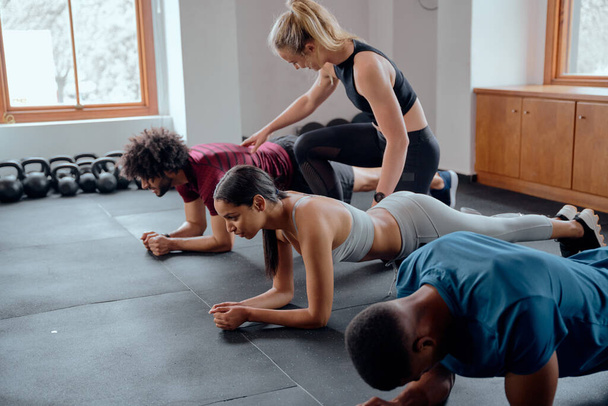 Trainer assisting group of multiracial young adults in sportswear doing plank exercises at the gym - Foto, afbeelding
