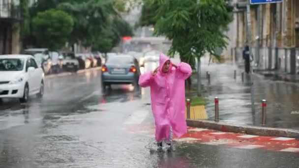 Jonge lachende vrouw met een roze regenjas op straat terwijl ze geniet van een wandeling door de stad op een regenachtige dag. - Video