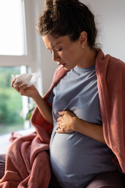 Беременная женщина, покрытая одеялом, сидит на кровати и чихает в ткань. Концепция простуды или вируса в период беременности - Фото, изображение