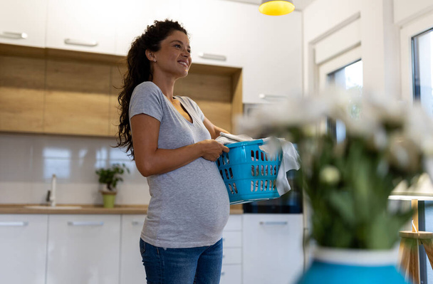 Όμορφη χαμογελαστή έγκυος γυναίκα μεταφέρουν καλάθι με πλυντήριο στο σπίτι - Φωτογραφία, εικόνα
