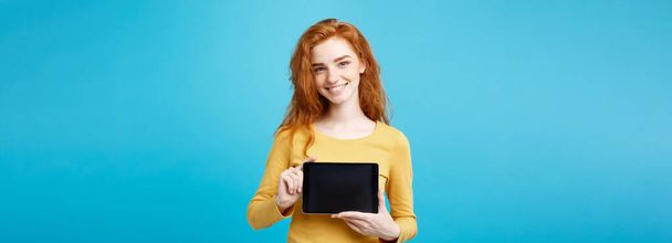 Бизнес - закройте портрет молодой красивой рыжеволосой девушки, улыбающейся, показывая цифровой экран планшета на черном. Синий пастельный фон
 - Фото, изображение