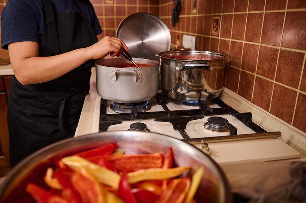 Close-up. Vista cortada de uma dona de casa usando concha, mexendo o suco de tomate fervente em uma panela, preparando o lecho de salada com tomates orgânicos e pimentas. Conserva pimentão doce de inverno - Foto, Imagem