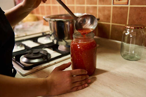 Close-up. Chef usando uma concha, derrama suco fervente de tomates suculentos orgânicos em uma lata de conserva esterilizada. Preparação de molho de tomate ou passata. Preservar legumes caseiros para o inverno. Alimentos enlatados - Foto, Imagem