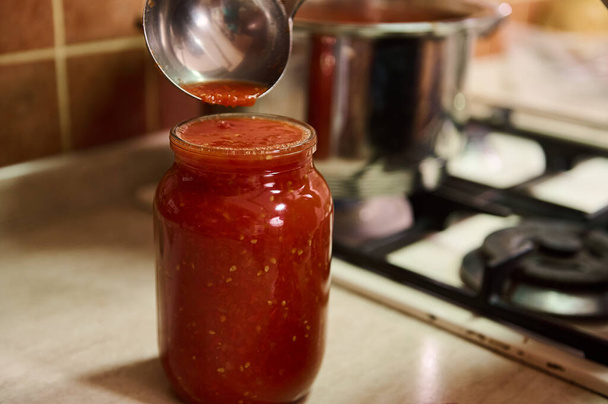 選択的フォーカス。滅菌された缶詰はキッチンカウンターの上に置くことができ、シェフはキッチンスプーンを使用して、鍋から新鮮な醸造トマトジュースを注ぎ、有機トマトから自家製パスタを調理します - 写真・画像