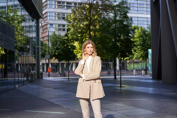 Hermosa mujer de negocios en traje beige, de pie cerca de edificios de oficinas en la calle, posando con confianza, expresión de la cara relajada. - Foto, imagen