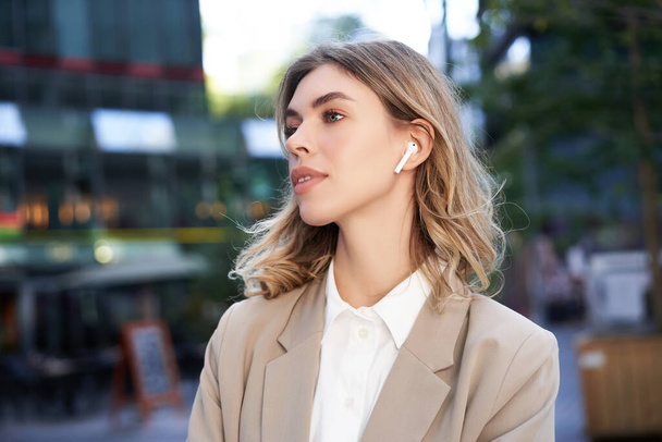 Close up portrait of blond businesswoman, confident corporate woman in headphones and beige suit, posing outdoors on street of city center. - Photo, Image