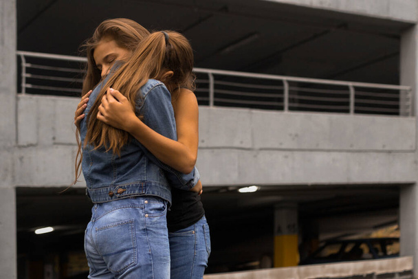 Moda portret dwóch stylowych nastolatek dziewczyny najlepsze przyjaciółki noszenie dżinsy przytula na zewnątrz w pobliżu nowoczesnego betonu budynku - Zdjęcie, obraz