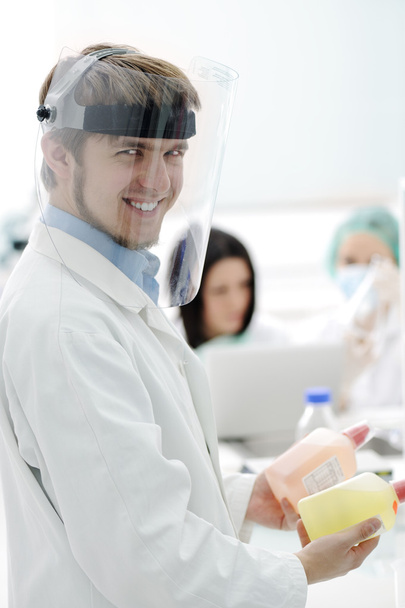 Young medical worker with glass mask working with his team in lab - Фото, изображение