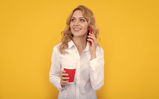 femme blonde positive avec tasse de café parlant au téléphone sur fond jaune. - Photo, image