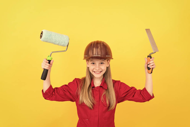 felice adolescente bambino in costruttore cappello duro con rullo di vernice e spatola sulla parete gialla. - Foto, immagini